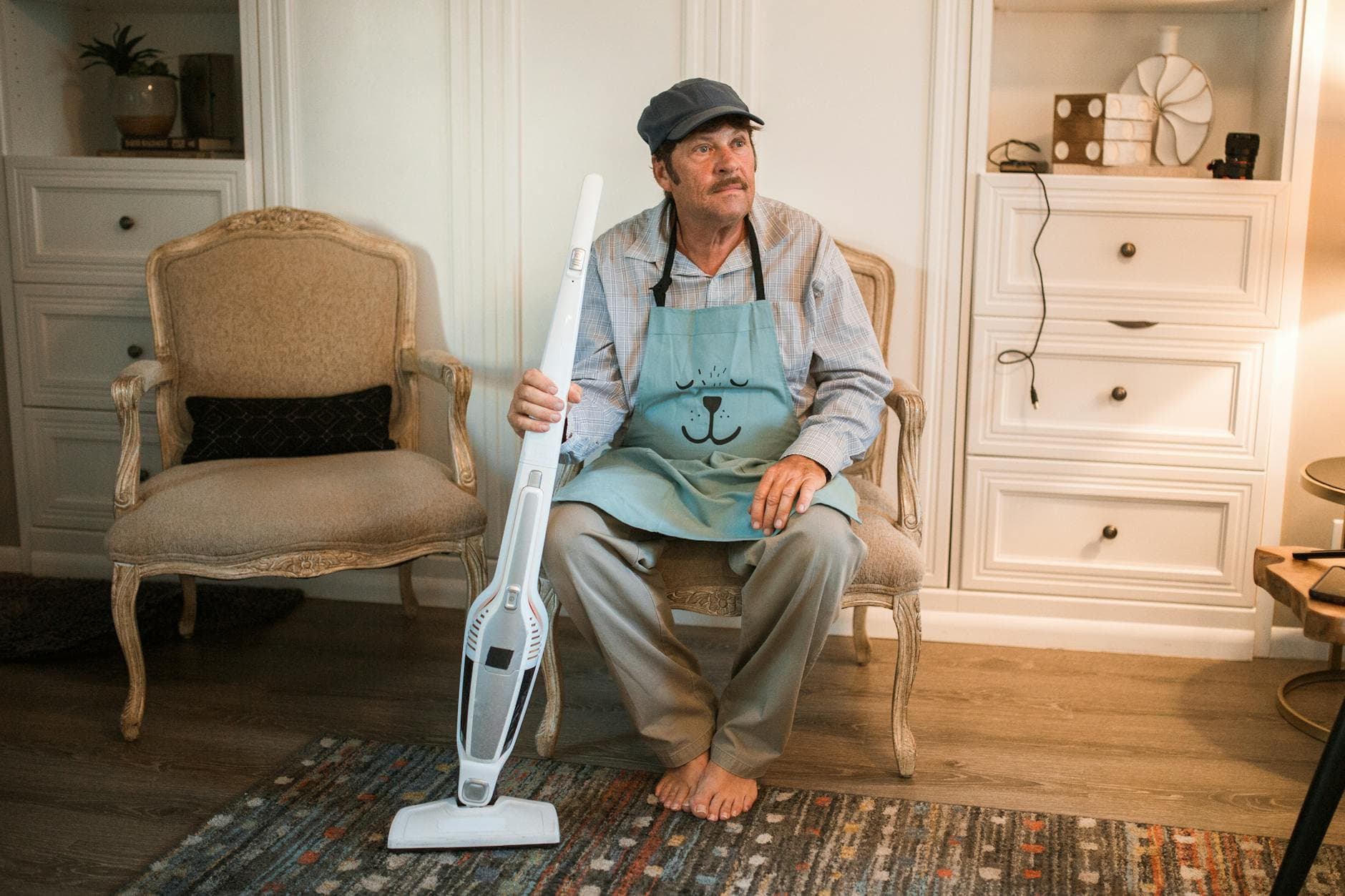 An adult man in a cap and apron holding a vacuum cleaner, sitting on an ornate chair in a cozy living room.
