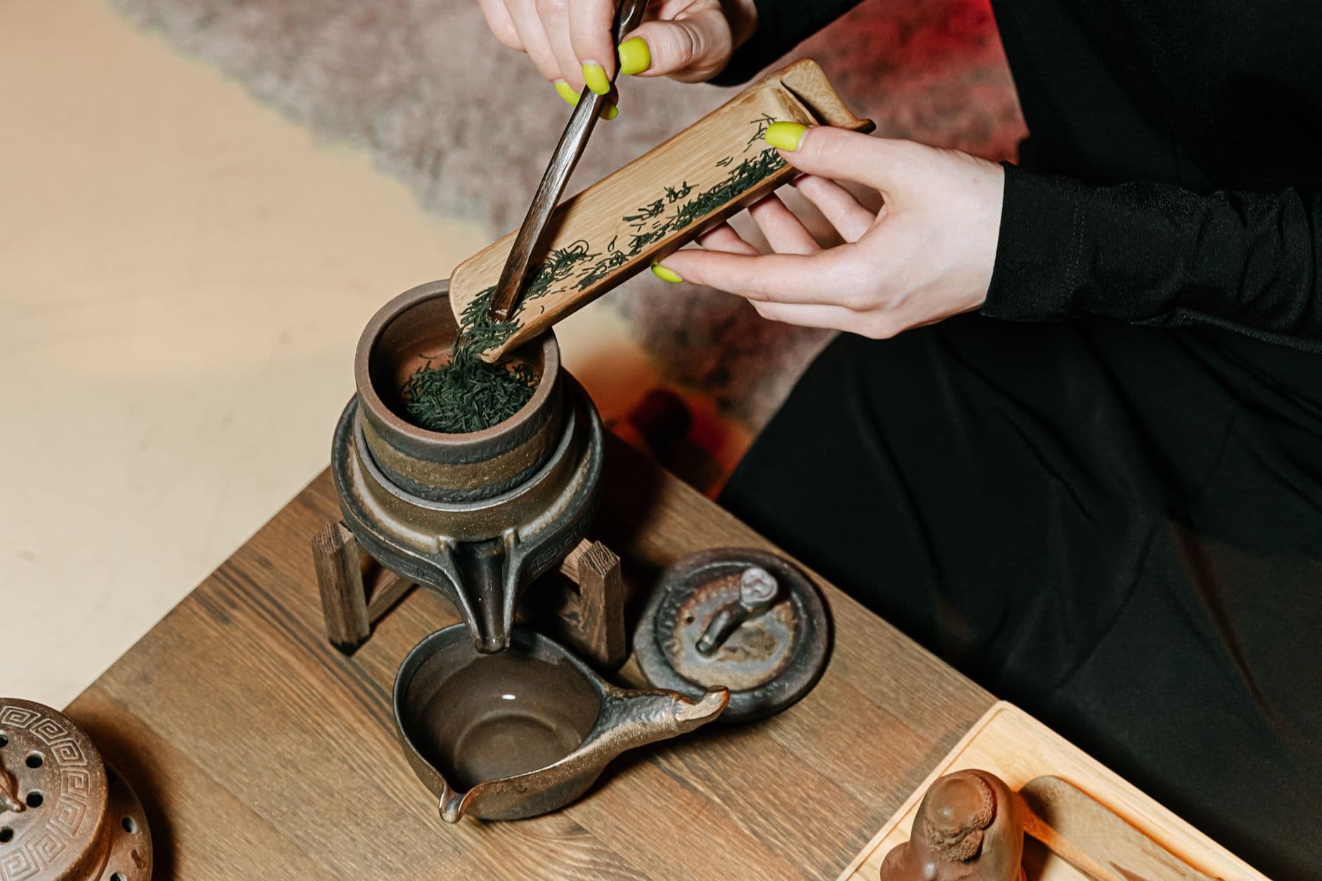 A person prepares tea by adding leaves with a bamboo scoop into a vintage teapot.